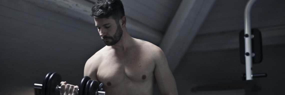 From below of muscular shirtless athletic male sitting on bench and doing bicep curls with dumbbell during intense training in dark studio