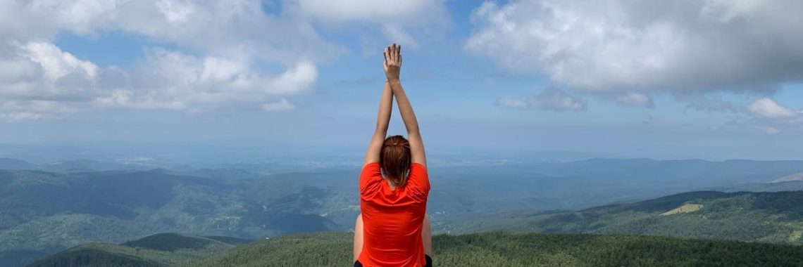 A woman in a red shirt relaxes on a mountain peak with arms raised, enjoying the scenic view.